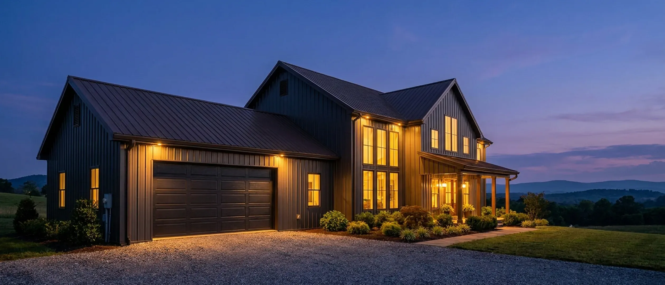 A barndominium at dusk with warm light glowing from every window and exterior lighting along the porch and roofline against a deep blue twilight sky.