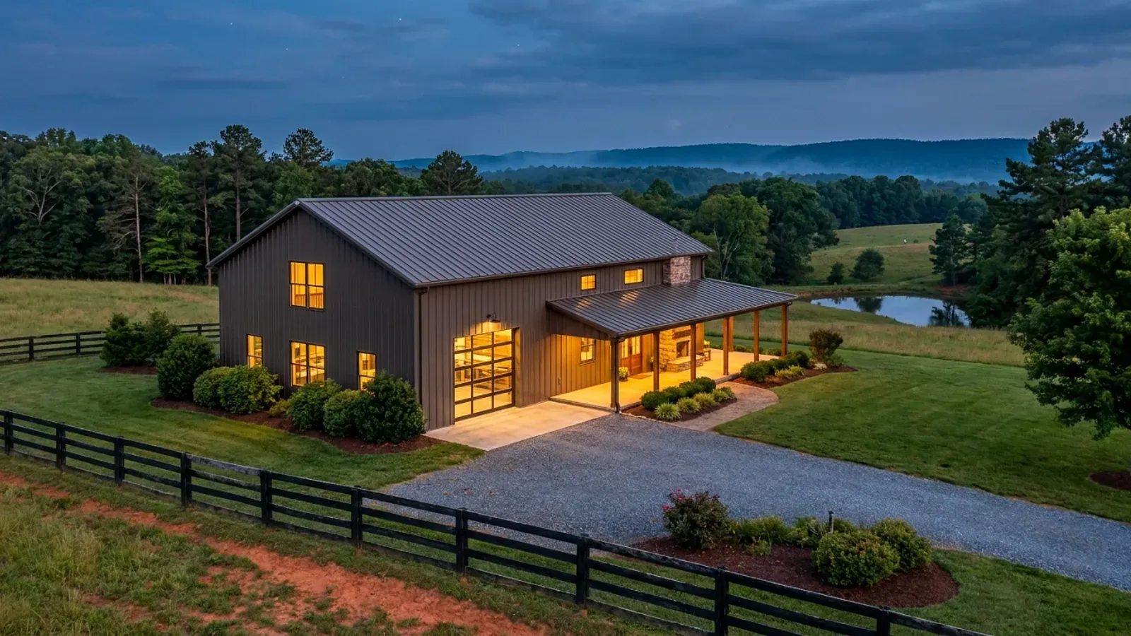A finished barndominium at dusk with warm interior lighting glowing through its large windows.